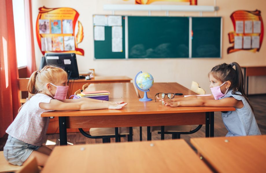 Two schoolgirls in medical masks are sitting at a school desk, opposite each other, group session
